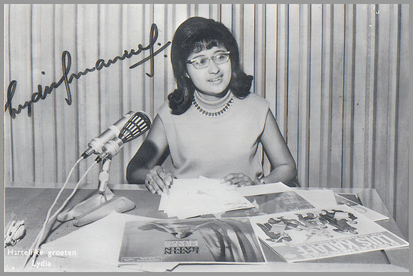 Lydia Emanuels in de studio van de Wereldomroep. Voor haar op tafel ligt een aantal lp’s, waaronder Latin Satin van Perez Prado en een lp van Luis Kalaf. Dit is een gesigneerde publiciteitsfoto die luisteraars bij de Wereldomroep konden bestellen. 